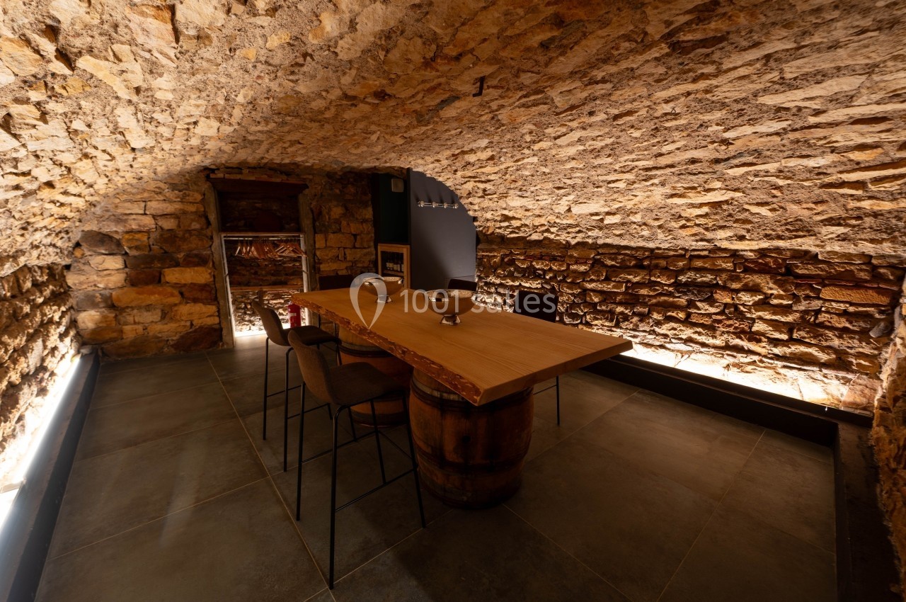 Salle voûtée en pierre avec une table en bois sur un tonneau, entourée de chaises, éclairage tamisé.