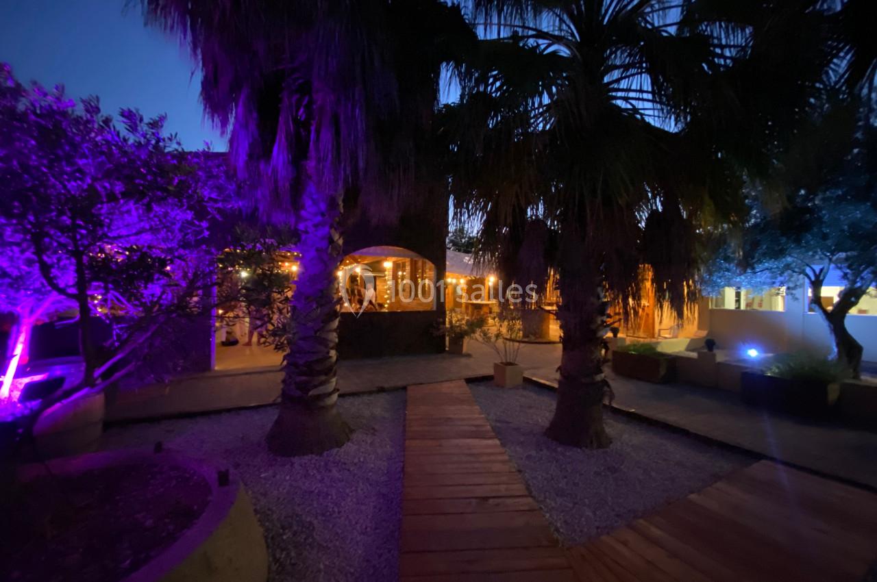 Allée en bois bordée de palmiers et éclairée par des lumières violettes et chaudes dans un cadre nocturne.