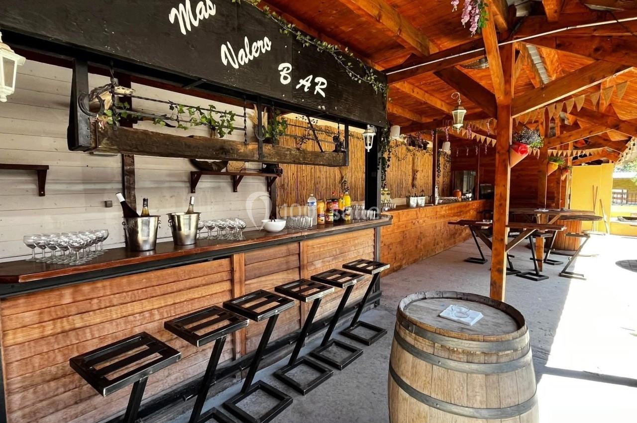 Bar extérieur en bois avec tabourets, tonneau décoratif, étagères à bouteilles et guirlandes suspendues sous un toit en bois.