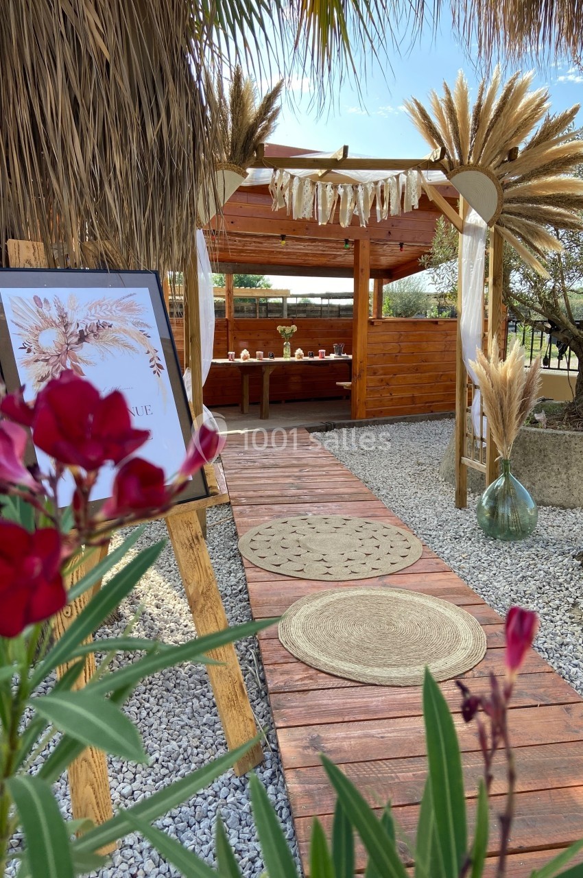 Allée en bois décorée de tapis ronds menant à une pergola ornée de pampas, avec fleurs rouges au premier plan.