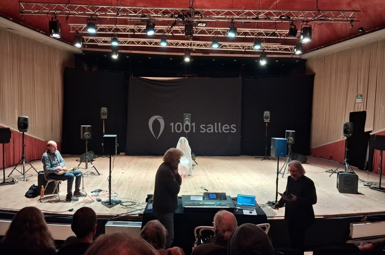 Une scène de théâtre avec des spectateurs, un technicien au premier plan et une personne voilée au centre de la scène.