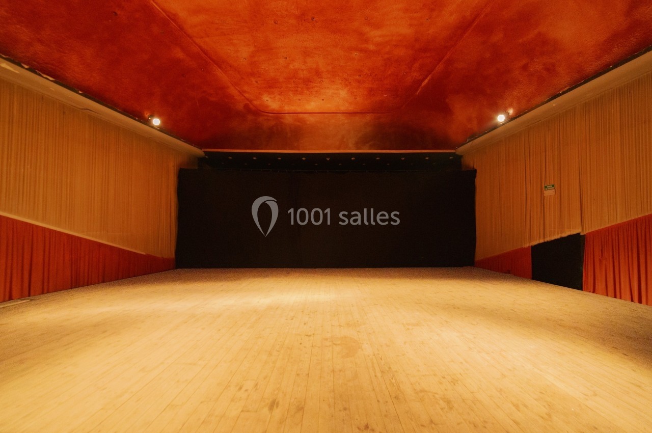 Salle de spectacle vide avec scène en bois, rideaux rouges sur les côtés et plafond de couleur orange.
