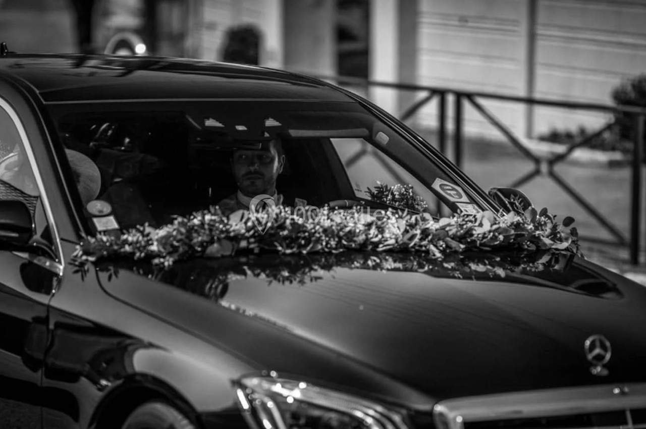 Voiture noire décorée de fleurs à l'avant, conduite par un homme, dans un contexte urbain.