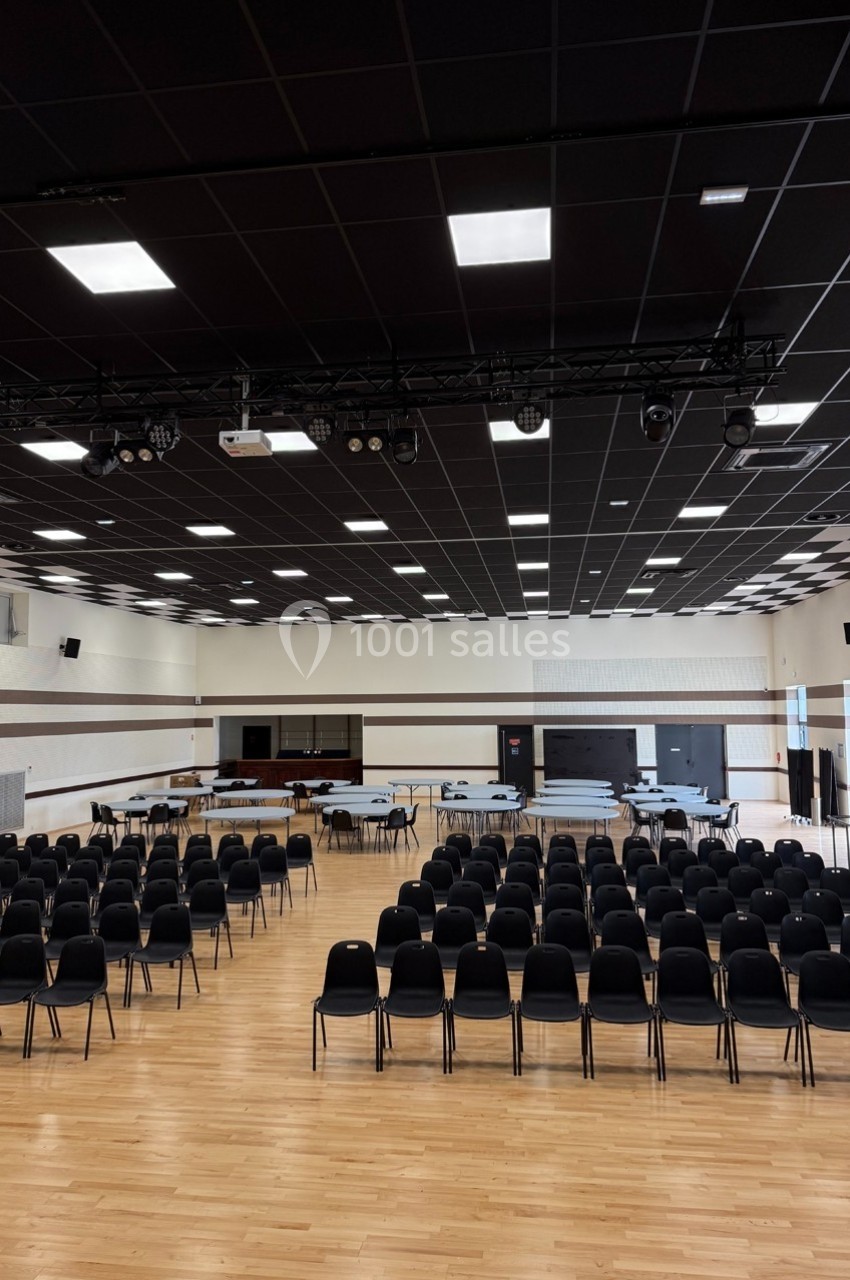 Salle polyvalente avec parquet, rangées de chaises noires, tables installées et plafond équipé de luminaires.