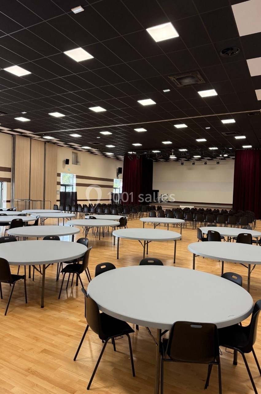 Salle polyvalente avec parquet, tables rondes, chaises noires, scène avec rideaux rouges et éclairage au plafond.