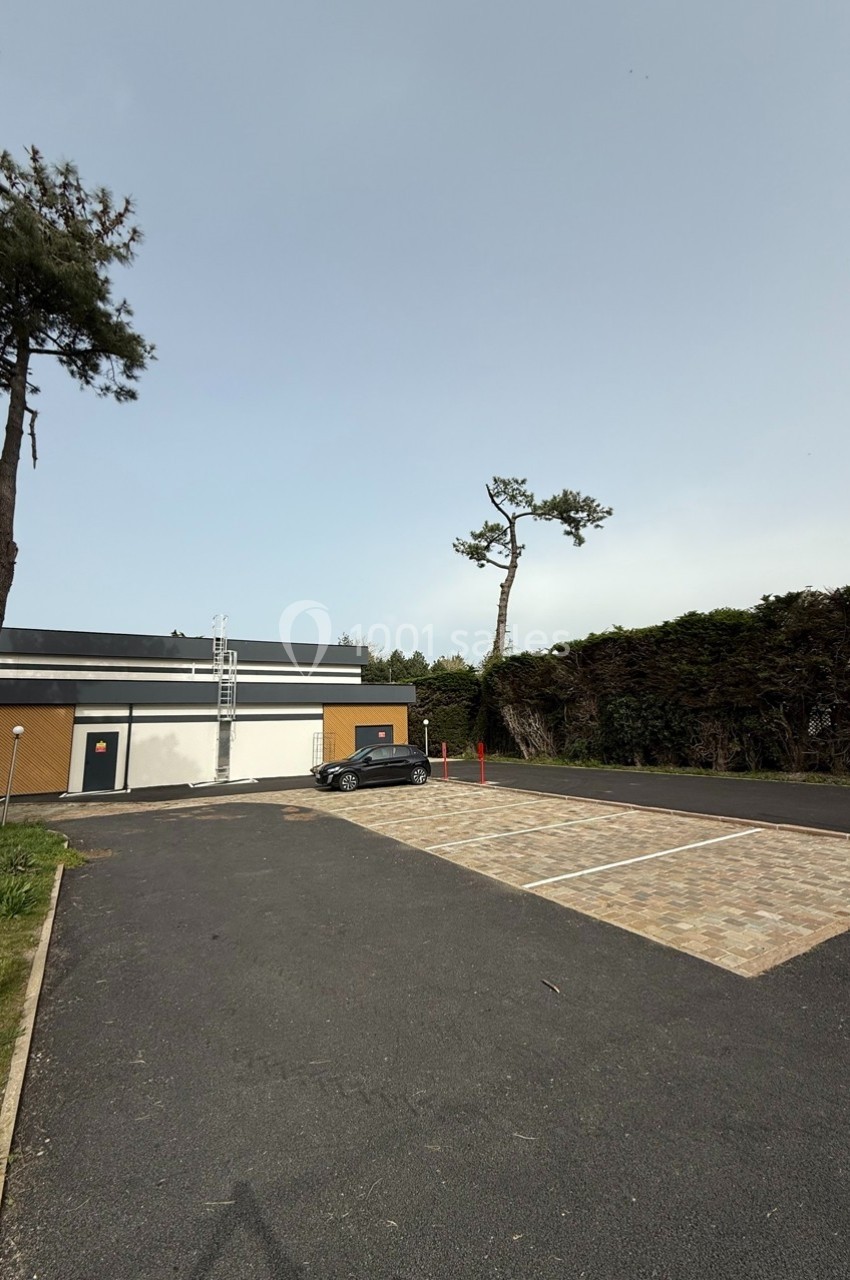 Parking vide avec quelques places pavées, une voiture stationnée, un bâtiment moderne et des arbres en arrière-plan.