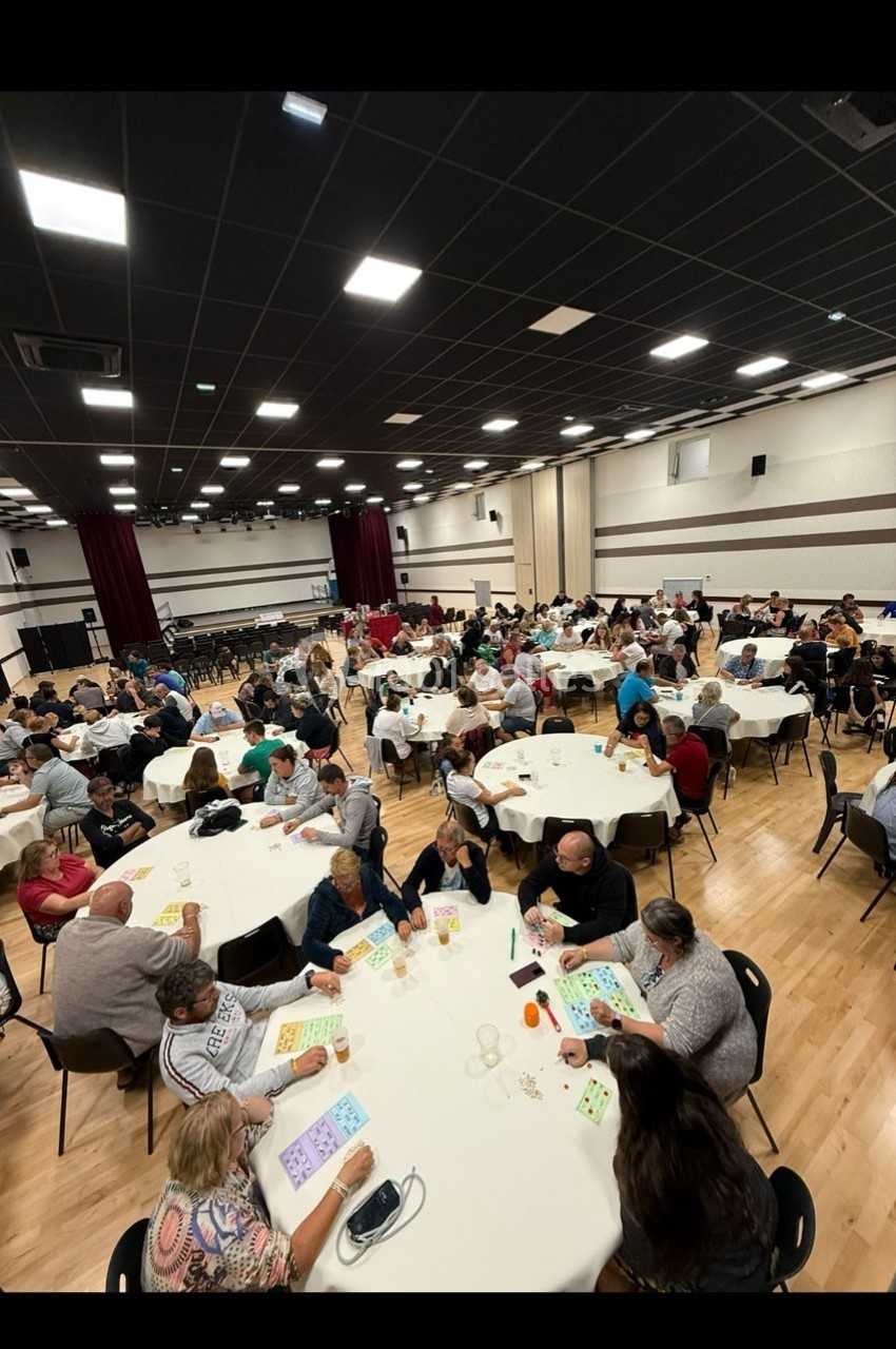 Des personnes participent à un loto dans une grande salle, assises à des tables rondes avec des cartons de jeu.