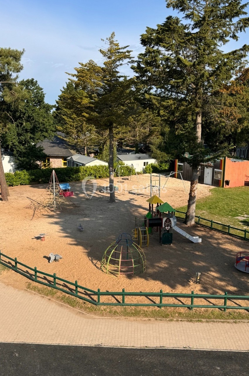 Aire de jeux extérieure avec toboggan, balançoires, structures d'escalade et sol en sable, entourée d'arbres.