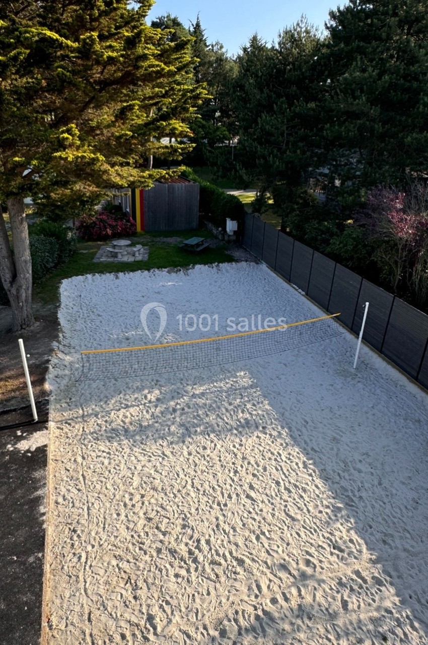 Terrain de sable avec un filet de sport, entouré d'arbres et de clôtures noires, sous un ciel dégagé.