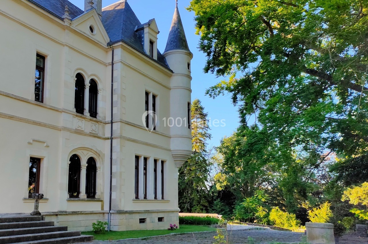Façade d'un château avec tourelle, entourée d'arbres et d'un jardin sous un ciel dégagé.