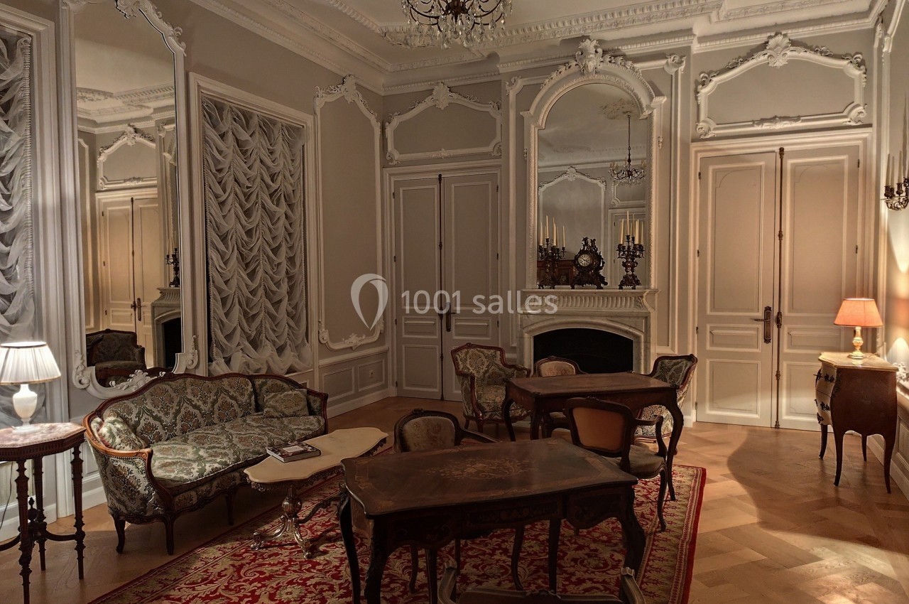 Salon élégant avec mobilier ancien, cheminée ornée, miroirs, lustre et éclairage tamisé sur tapis rouge.