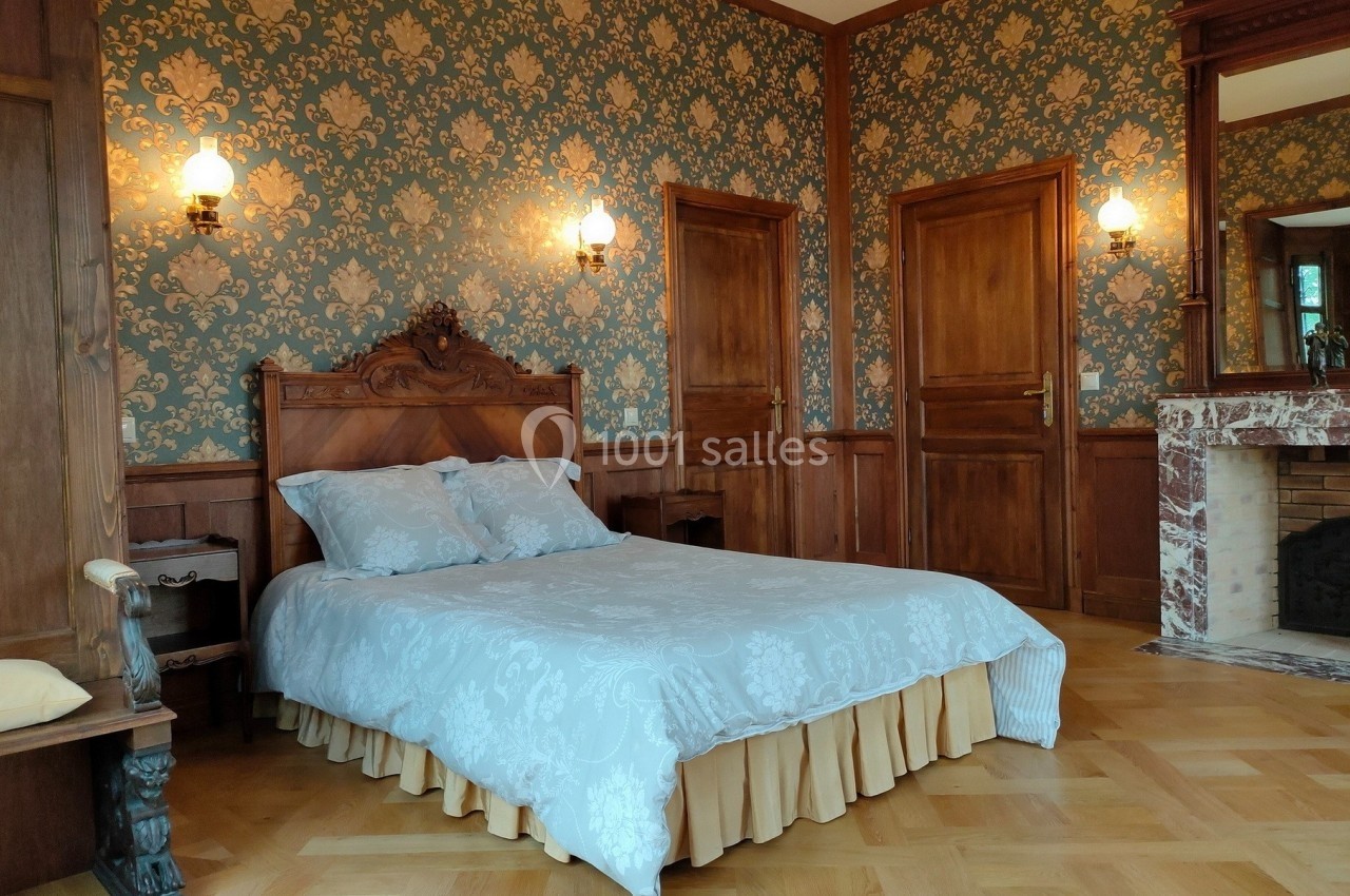 Chambre avec lit double, parquet en bois, murs ornés de papier peint à motifs et cheminée en marbre.
