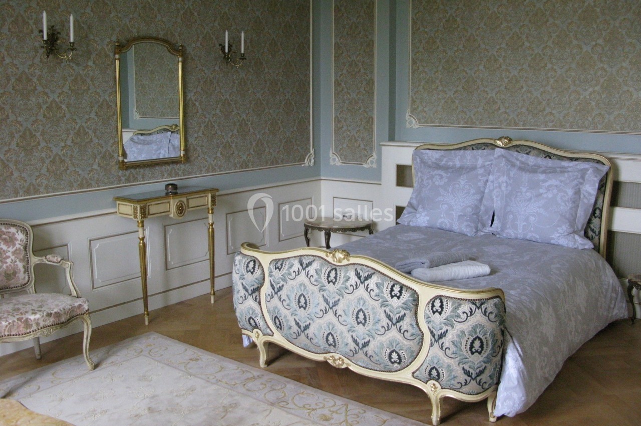 Chambre élégante avec lit à motifs floraux, miroir doré, fauteuil et murs ornés de papier peint classique.