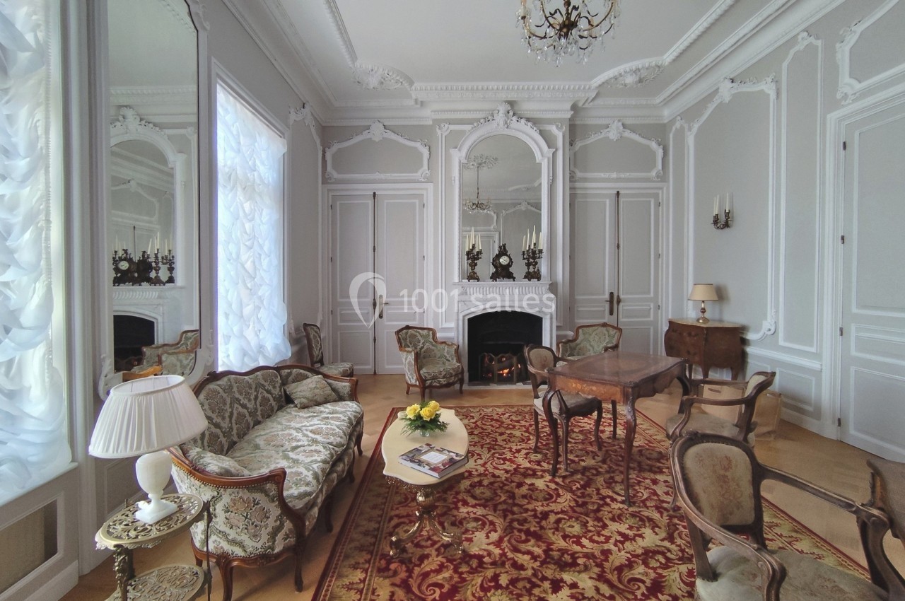 Salon élégant avec moulures, cheminée, meubles anciens, lustre et tapis rouge orné, baigné de lumière naturelle.