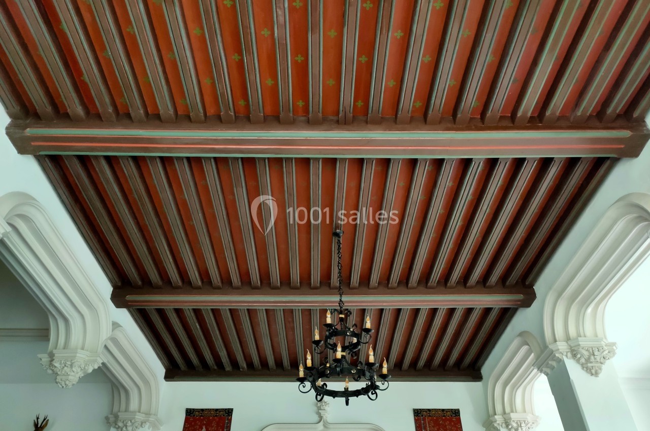 Plafond en bois à poutres apparentes avec un lustre en fer forgé suspendu, entouré d'arches blanches décoratives.