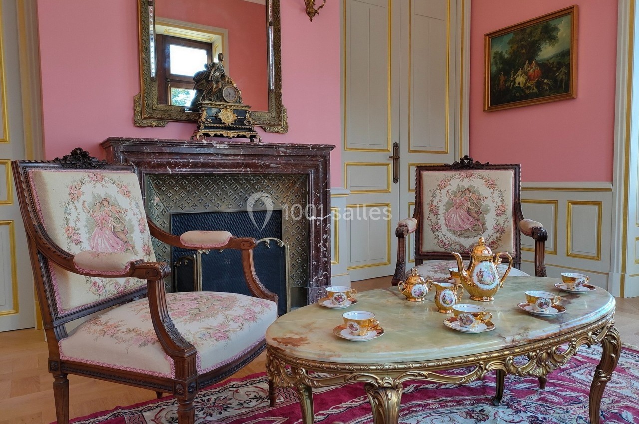Salon élégant avec murs roses, cheminée en marbre, meubles anciens et service à thé doré sur une table ornée.