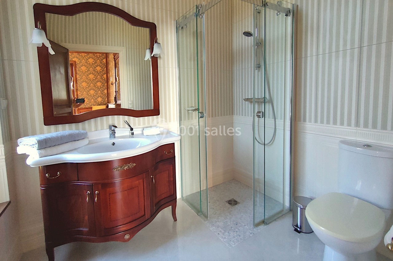Salle de bain avec meuble en bois, lavabo, miroir, douche vitrée et toilettes sur un sol clair.