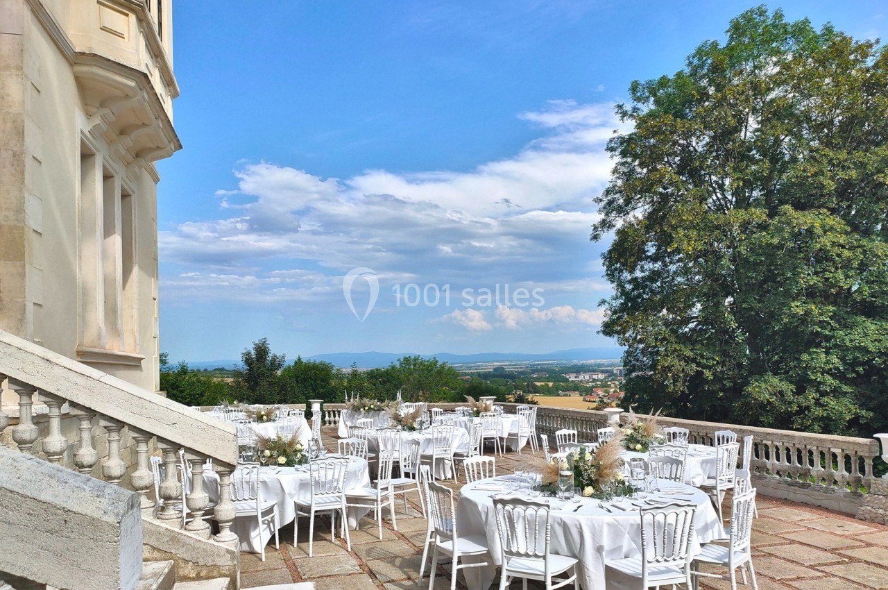 Tables dressées pour un événement en plein air sur une terrasse avec vue sur la campagne et un grand arbre.