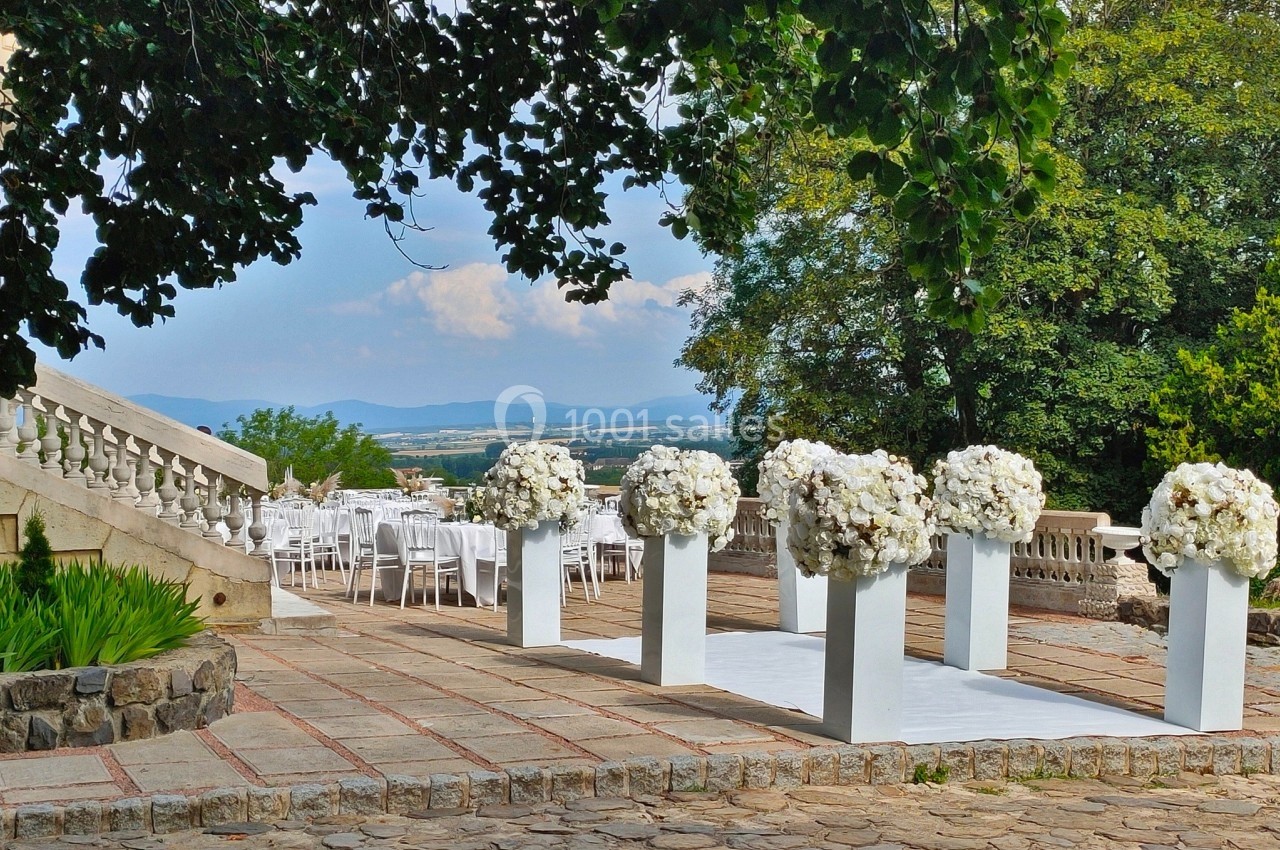 Allée décorée de colonnes blanches ornées de fleurs pour une cérémonie en plein air, avec des chaises en arrière-plan.