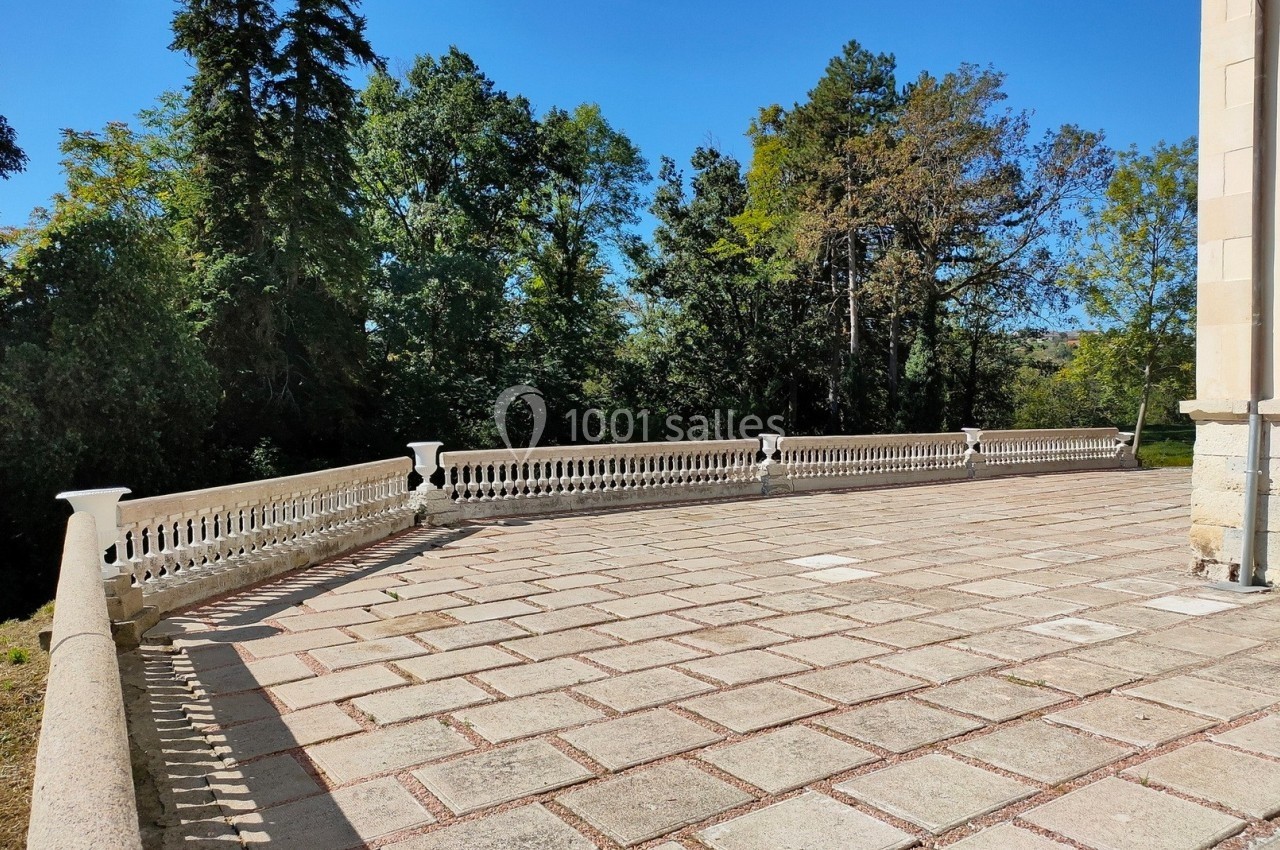Terrasse en pierre bordée de balustrades blanches, entourée d'arbres sous un ciel bleu clair.