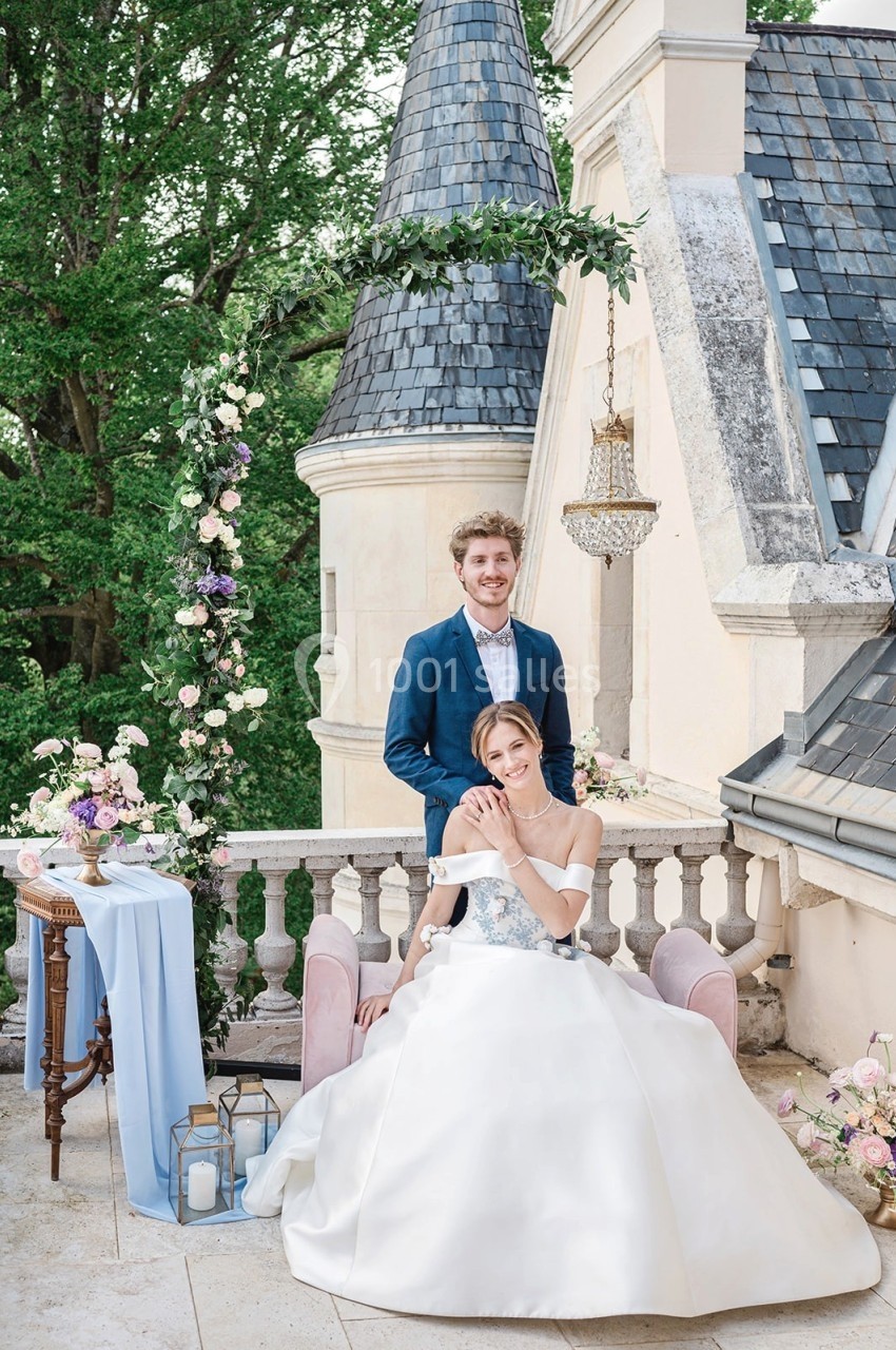 Un couple en tenue de mariage pose sur une terrasse élégante avec un décor floral et un château en arrière-plan.
