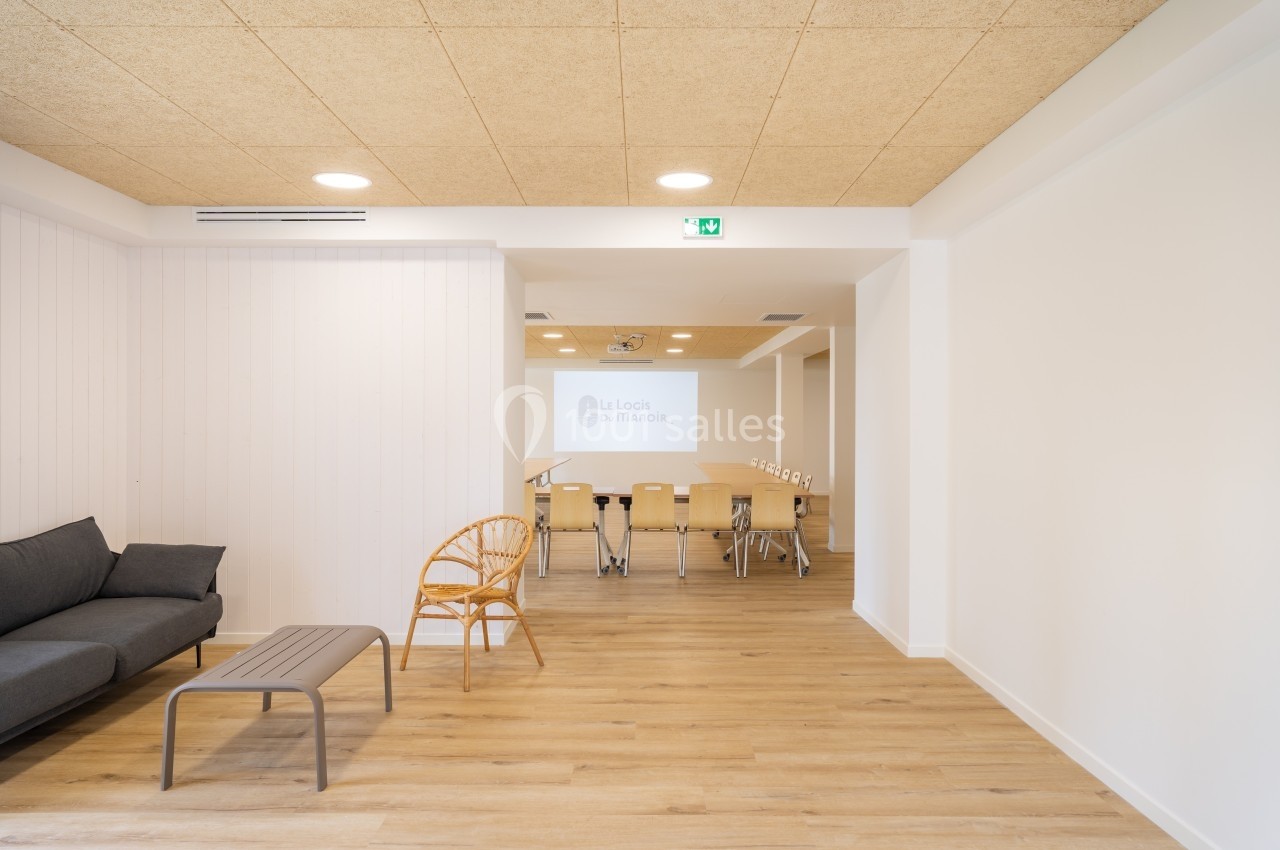 Salle lumineuse avec parquet, coin salon avec canapé et chaise en rotin, et espace de réunion avec table et chaises.