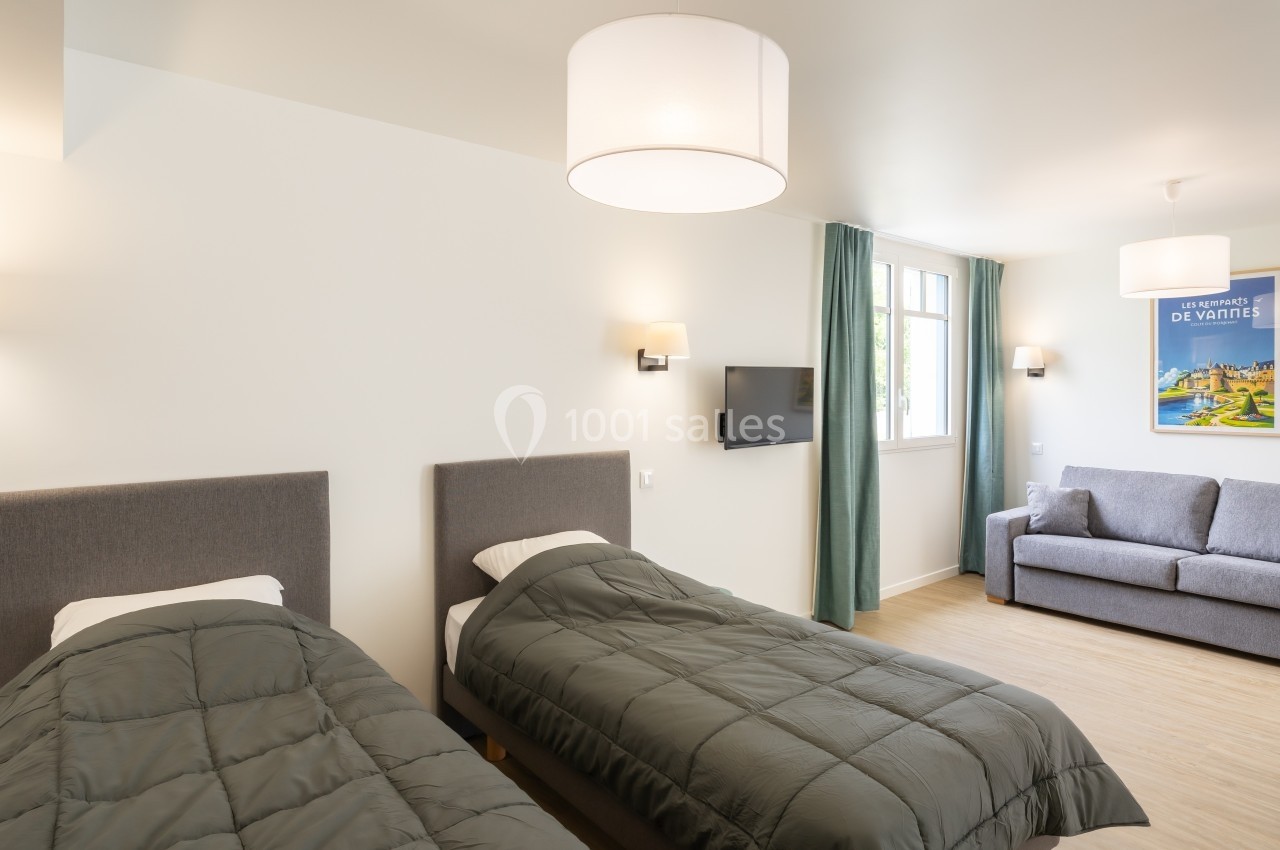 Chambre lumineuse avec deux lits simples, un canapé, une télévision murale et une grande fenêtre avec rideaux verts.
