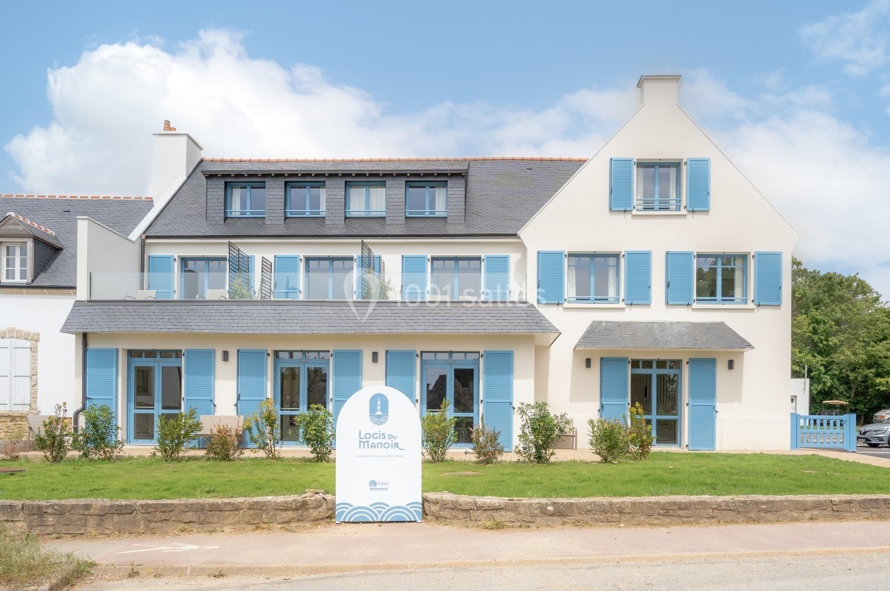 Façade d'un bâtiment moderne à deux étages avec volets bleus, entouré de végétation et ciel dégagé.