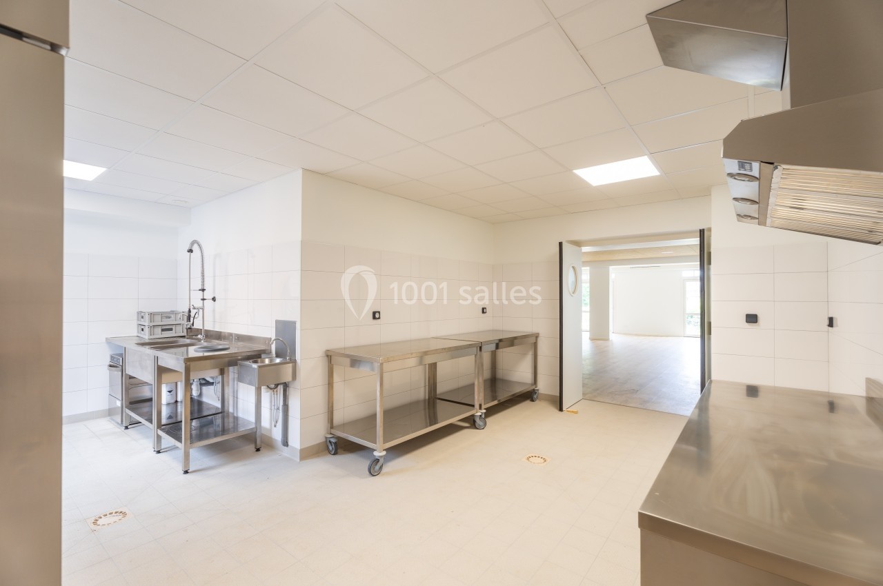 Cuisine professionnelle vide avec plans de travail en inox, évier, carrelage blanc et éclairage au plafond.