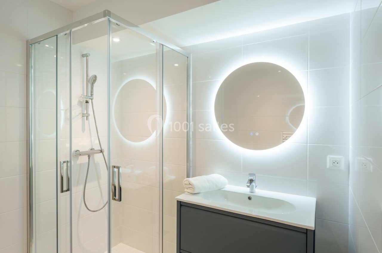 Salle de bain moderne avec douche vitrée, lavabo sur meuble noir et miroir rond éclairé.
