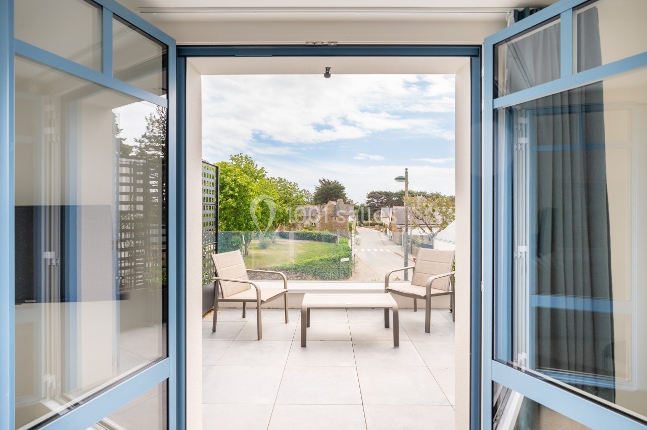Vue depuis une pièce sur une terrasse aménagée avec des fauteuils et une table, donnant sur un jardin et une rue.
