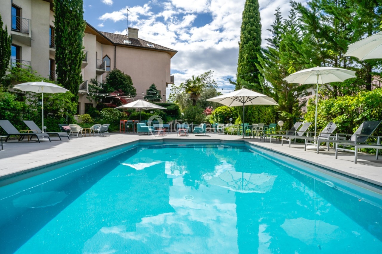 Piscine extérieure entourée de chaises longues, parasols et végétation, avec un bâtiment en arrière-plan.