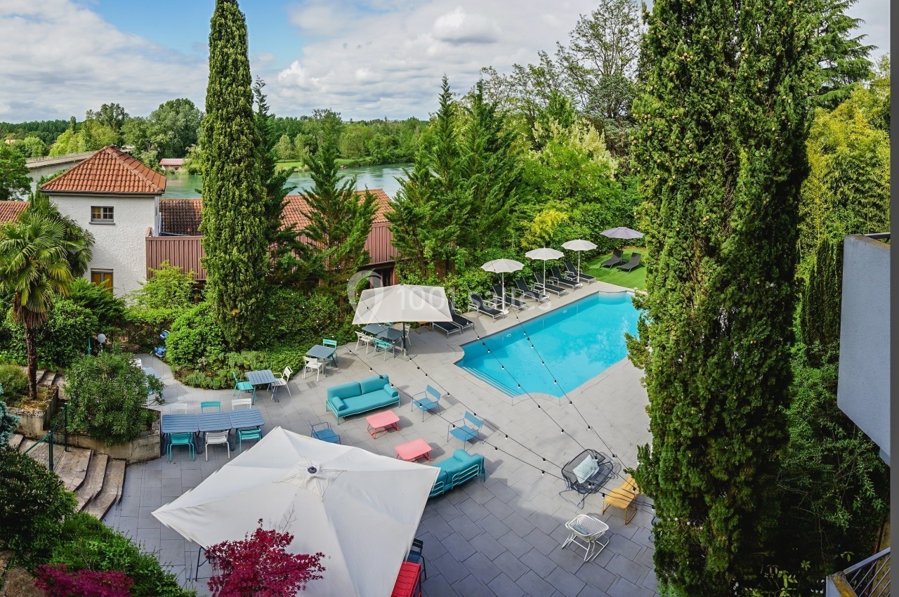 Vue aérienne d'une terrasse avec piscine, transats, parasols et végétation luxuriante entourant des bâtiments.