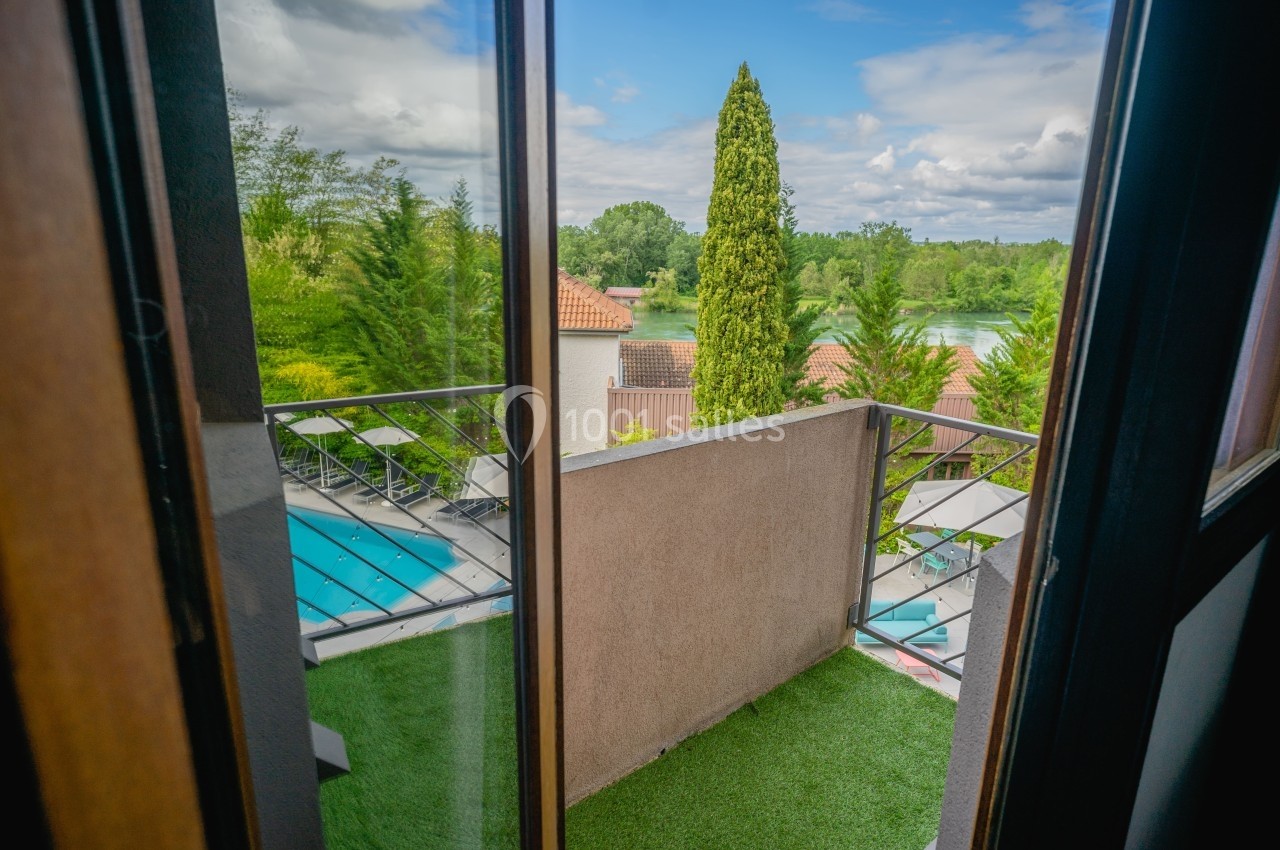 Vue depuis une porte-fenêtre donnant sur un petit balcon avec gazon artificiel, piscine et paysage verdoyant.