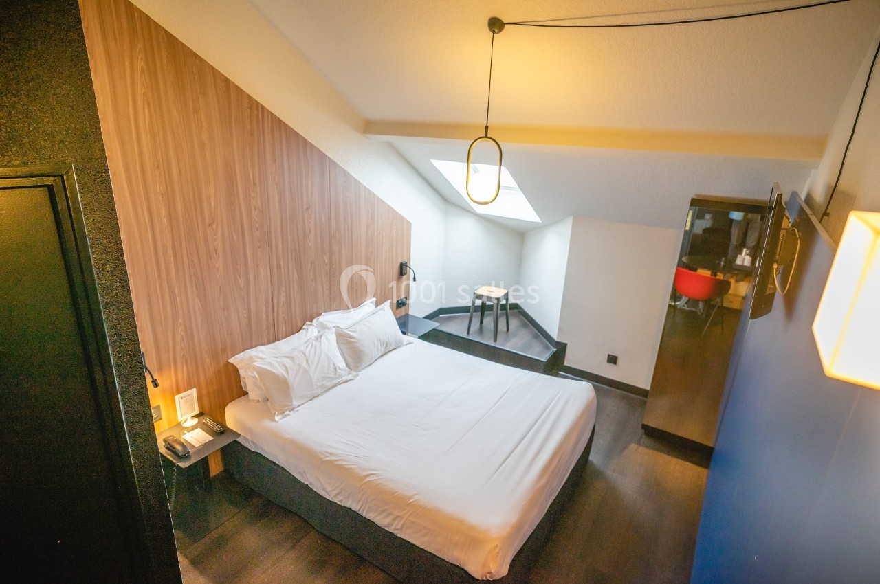 Chambre mansardée avec lit double, mur en bois, bureau, chaise et lumière naturelle provenant d'une fenêtre de toit.