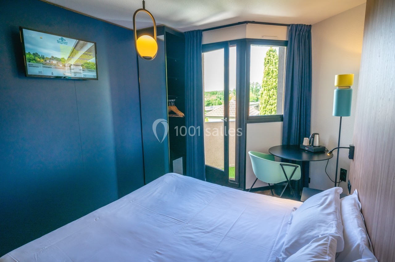 Chambre d'hôtel avec lit double, bureau, chaise, télévision murale et fenêtre donnant sur un paysage verdoyant.