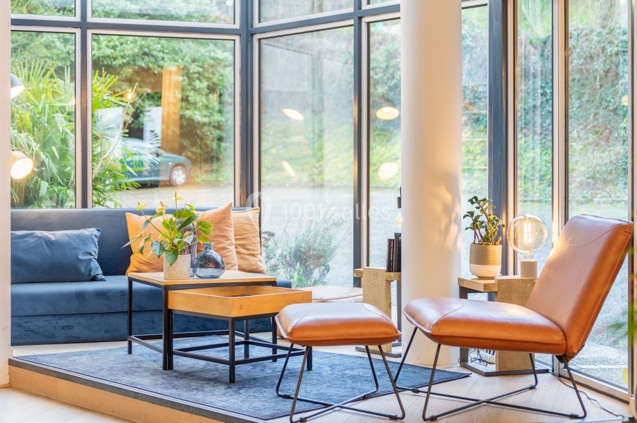 Salon lumineux avec fauteuils en cuir, canapé bleu, table basse en bois et grandes baies vitrées donnant sur un jardin.
