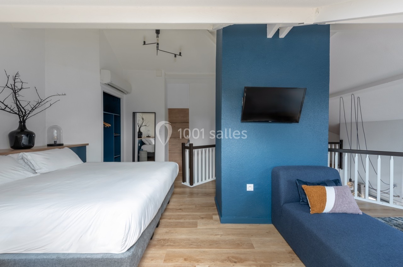 Chambre moderne avec lit double, canapé bleu, mur accent bleu, télévision murale et parquet clair.