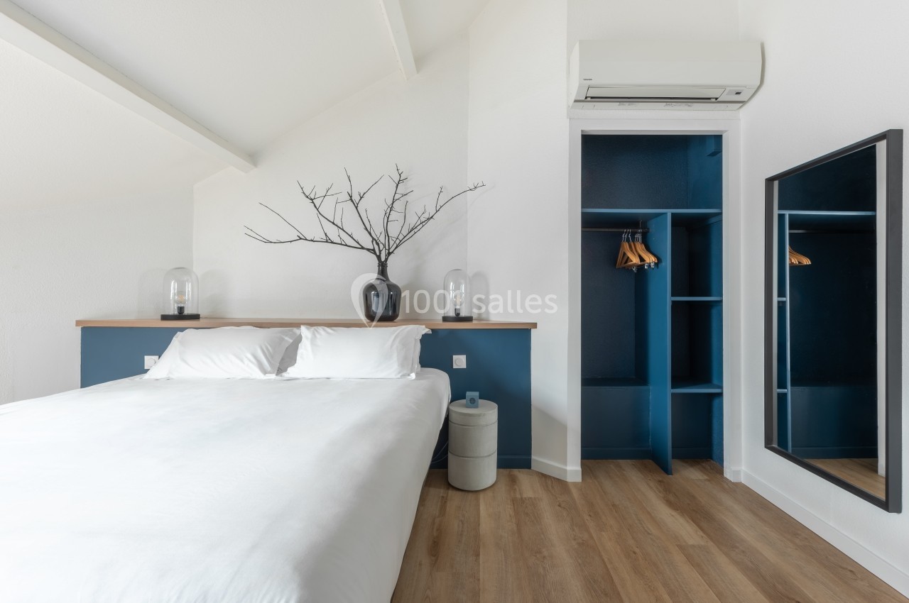 Chambre lumineuse avec lit double, étagère en bois, placard ouvert bleu, miroir et climatisation murale.