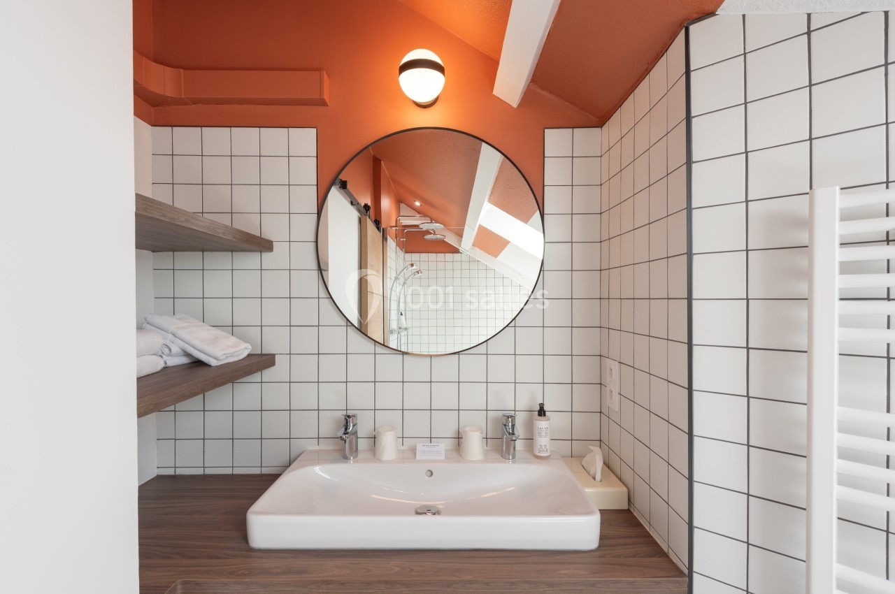 Lavabo blanc avec miroir rond, carrelage mural blanc, étagères en bois et mur orange sous un éclairage doux.