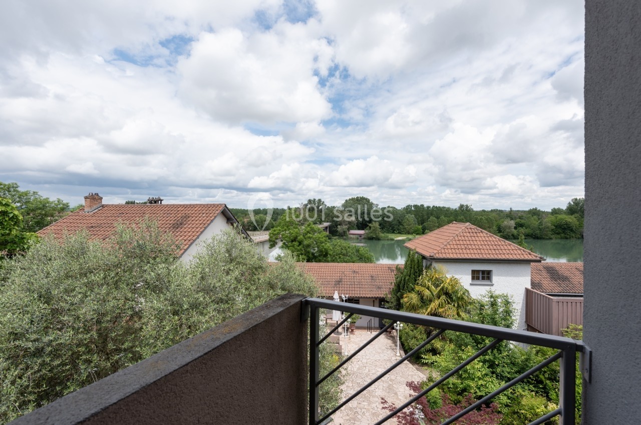 Vue depuis un balcon sur des toits de maisons, un jardin arboré et un plan d'eau entouré de végétation sous un ciel nuageux.
