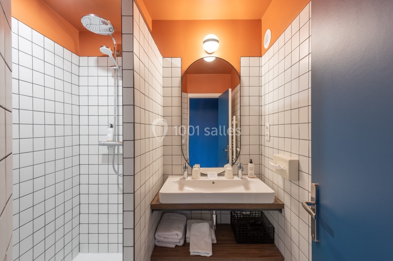 Salle de bain avec carrelage blanc, douche, lavabo avec miroir, serviettes pliées et mur orange vif.