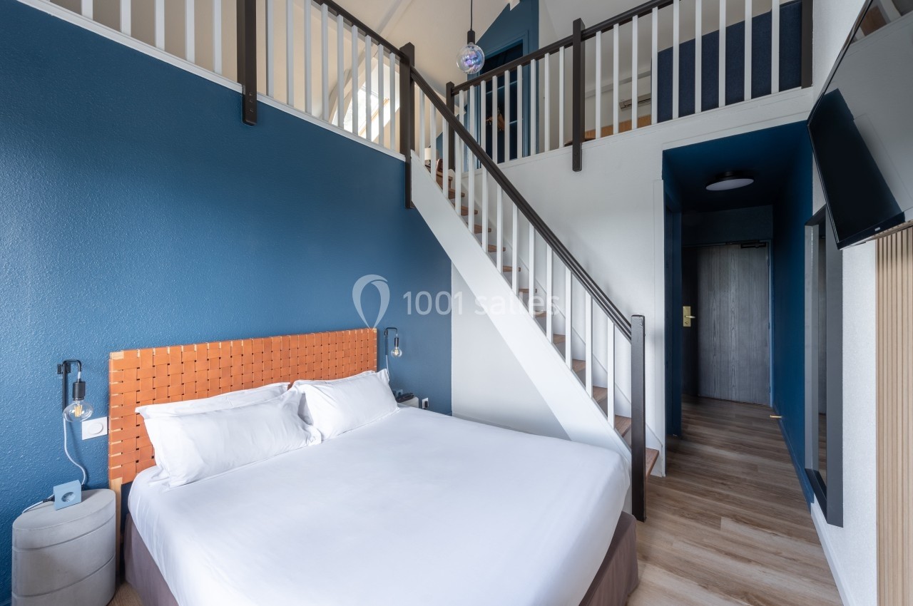 Chambre avec lit double, mezzanine, murs bleus, escalier en bois blanc et télévision fixée au mur.