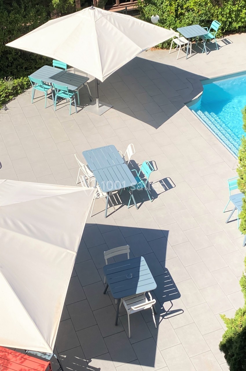 Terrasse avec tables, chaises, parasols blancs et piscine adjacente, entourée de verdure.