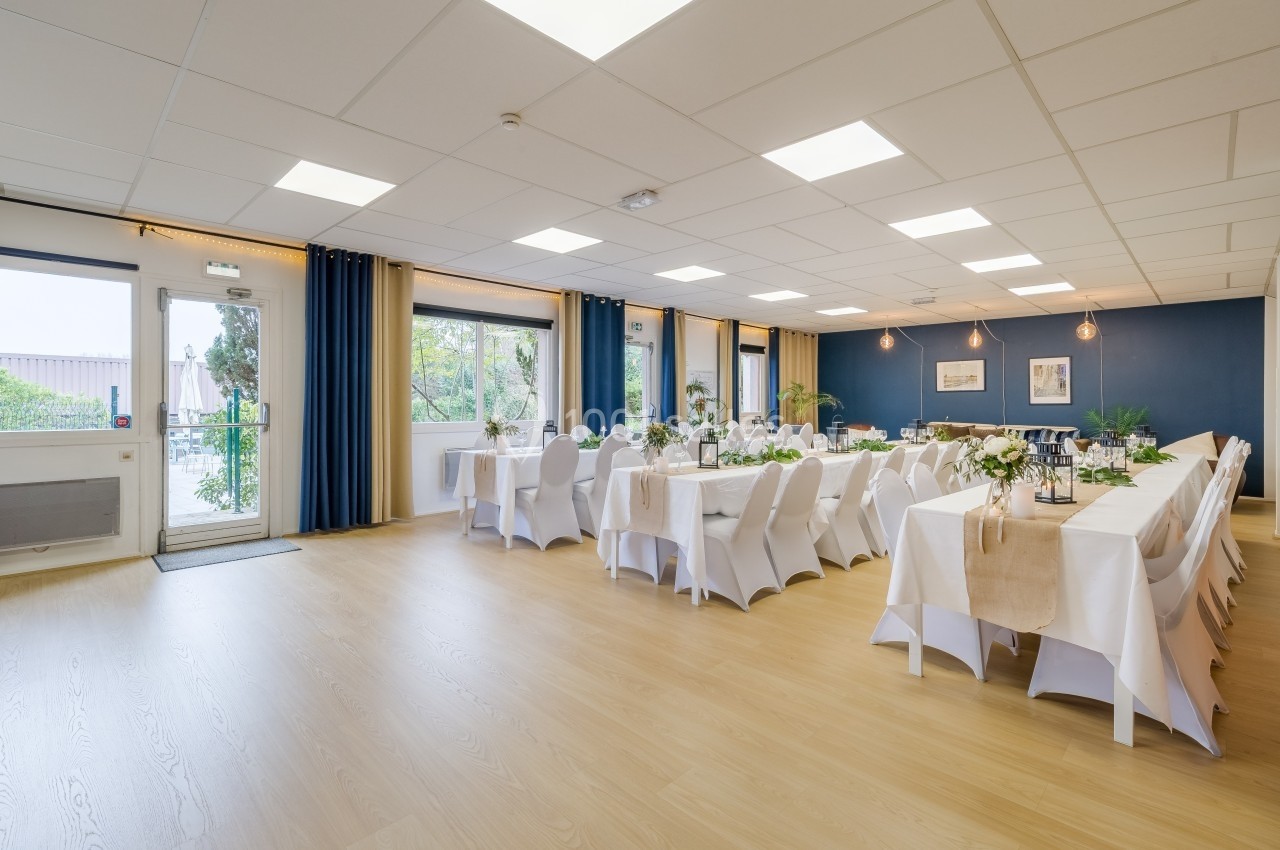 Salle lumineuse aménagée pour un événement, avec tables dressées, nappes blanches et décorations florales.