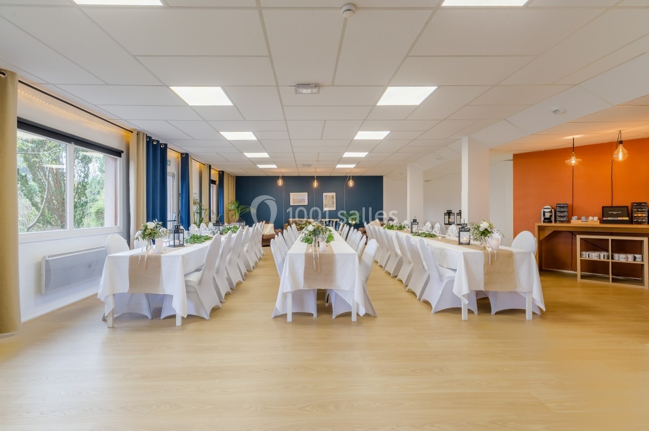 Salle lumineuse aménagée avec deux longues tables dressées pour un événement, décorée de fleurs et nappes blanches.