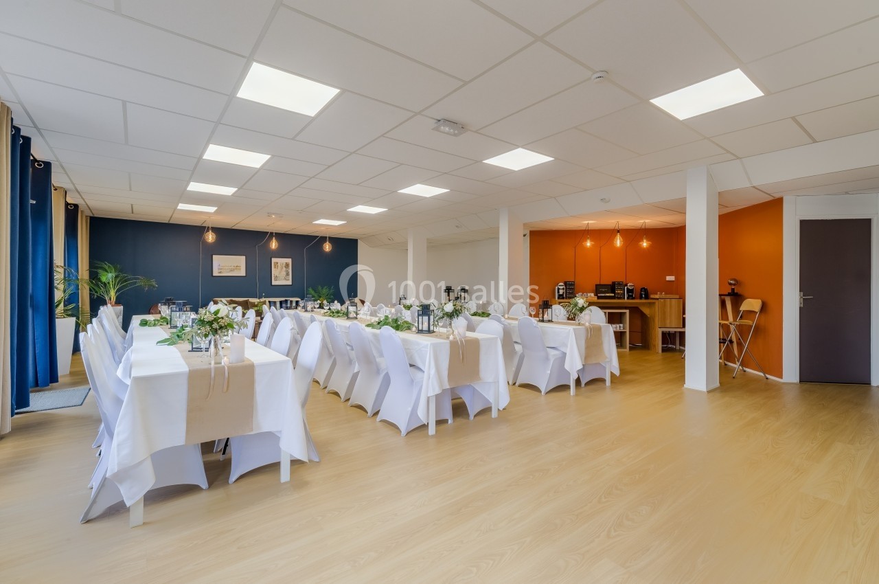 Salle lumineuse aménagée pour un repas, avec tables décorées, chaises blanches et un buffet en arrière-plan.
