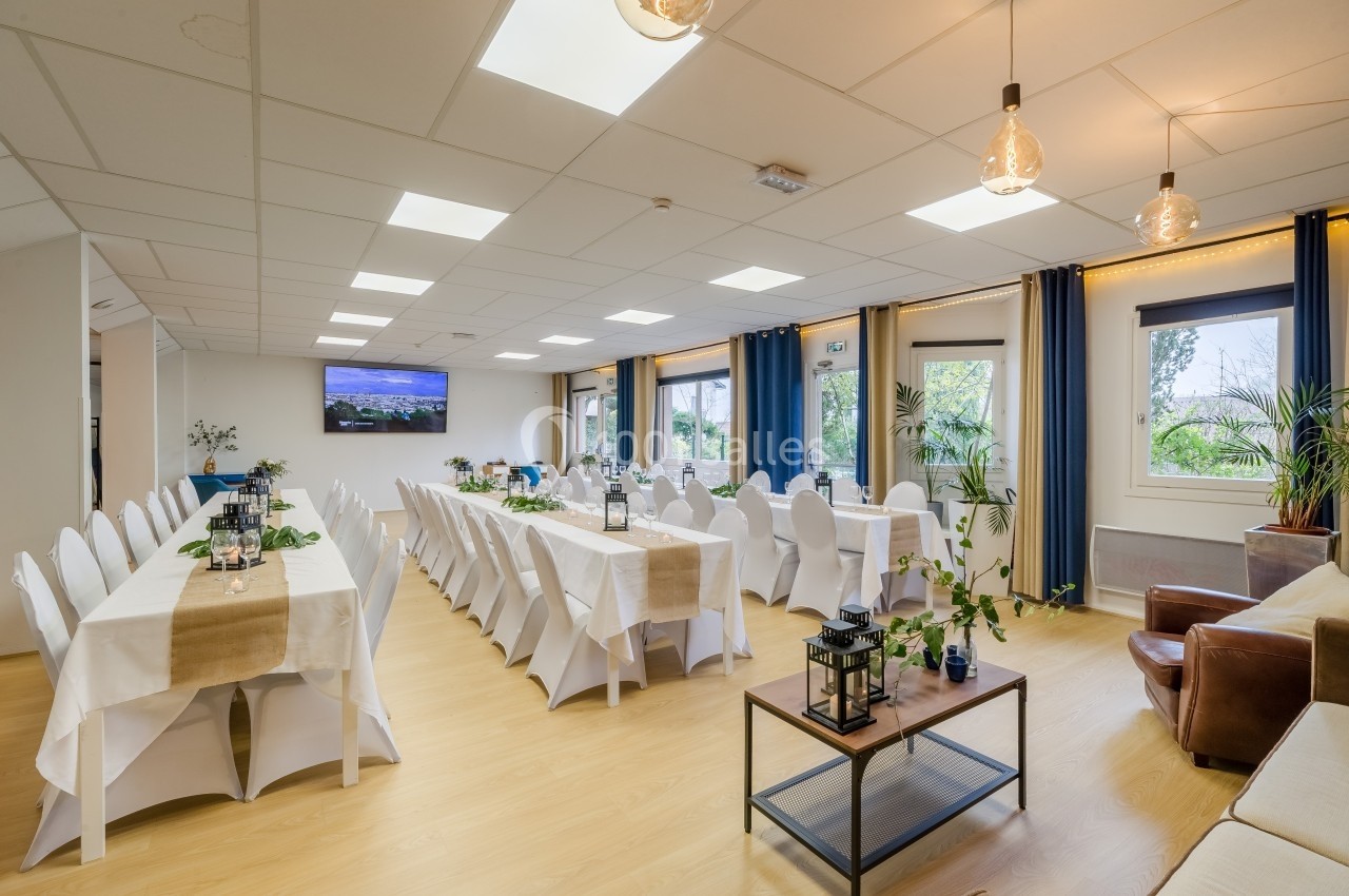 Salle lumineuse avec deux longues tables dressées, chaises blanches, plantes décoratives et grandes fenêtres donnant sur l…