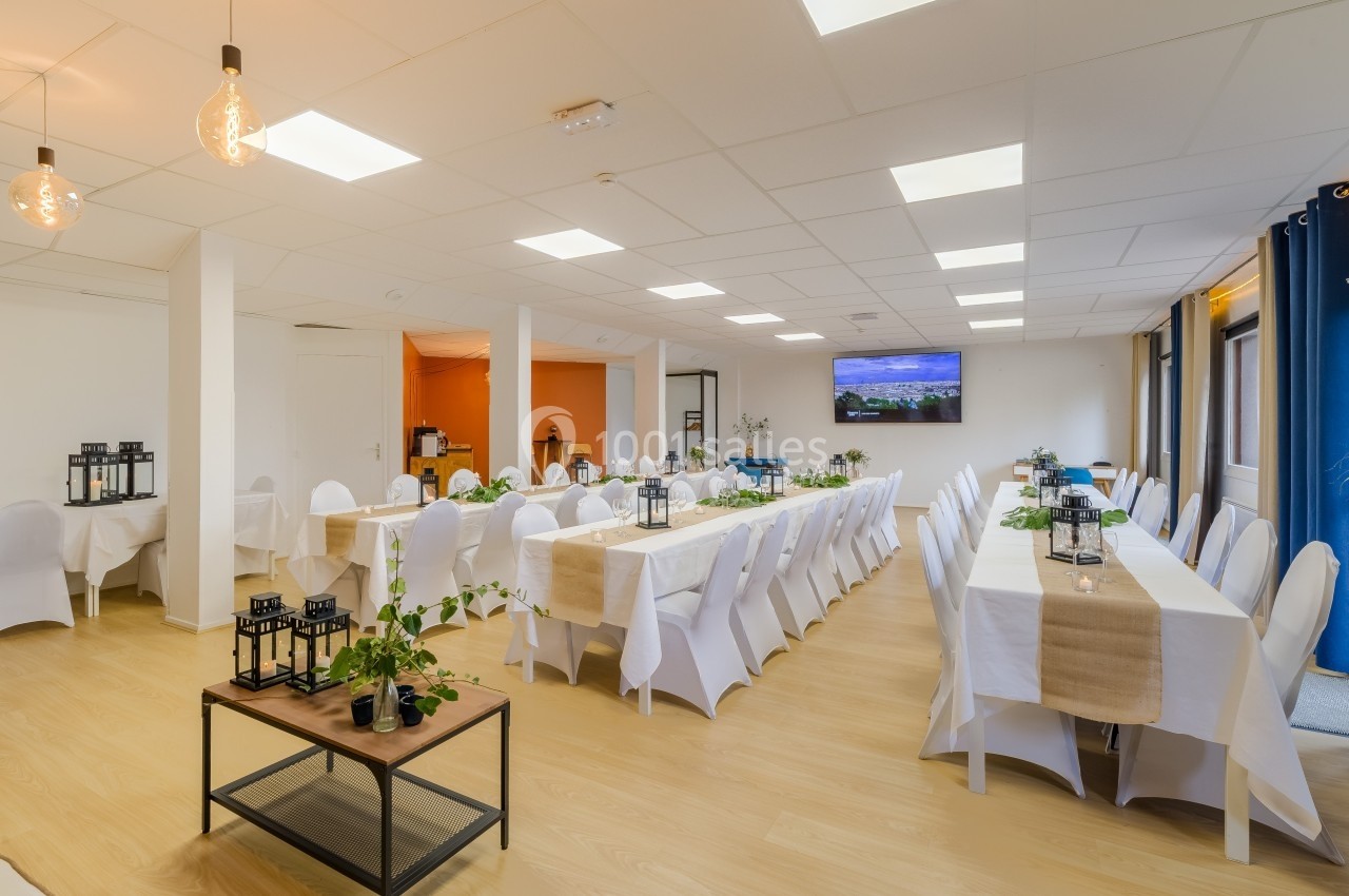 Salle lumineuse aménagée pour un événement, avec des tables décorées, des chaises blanches et un écran mural.