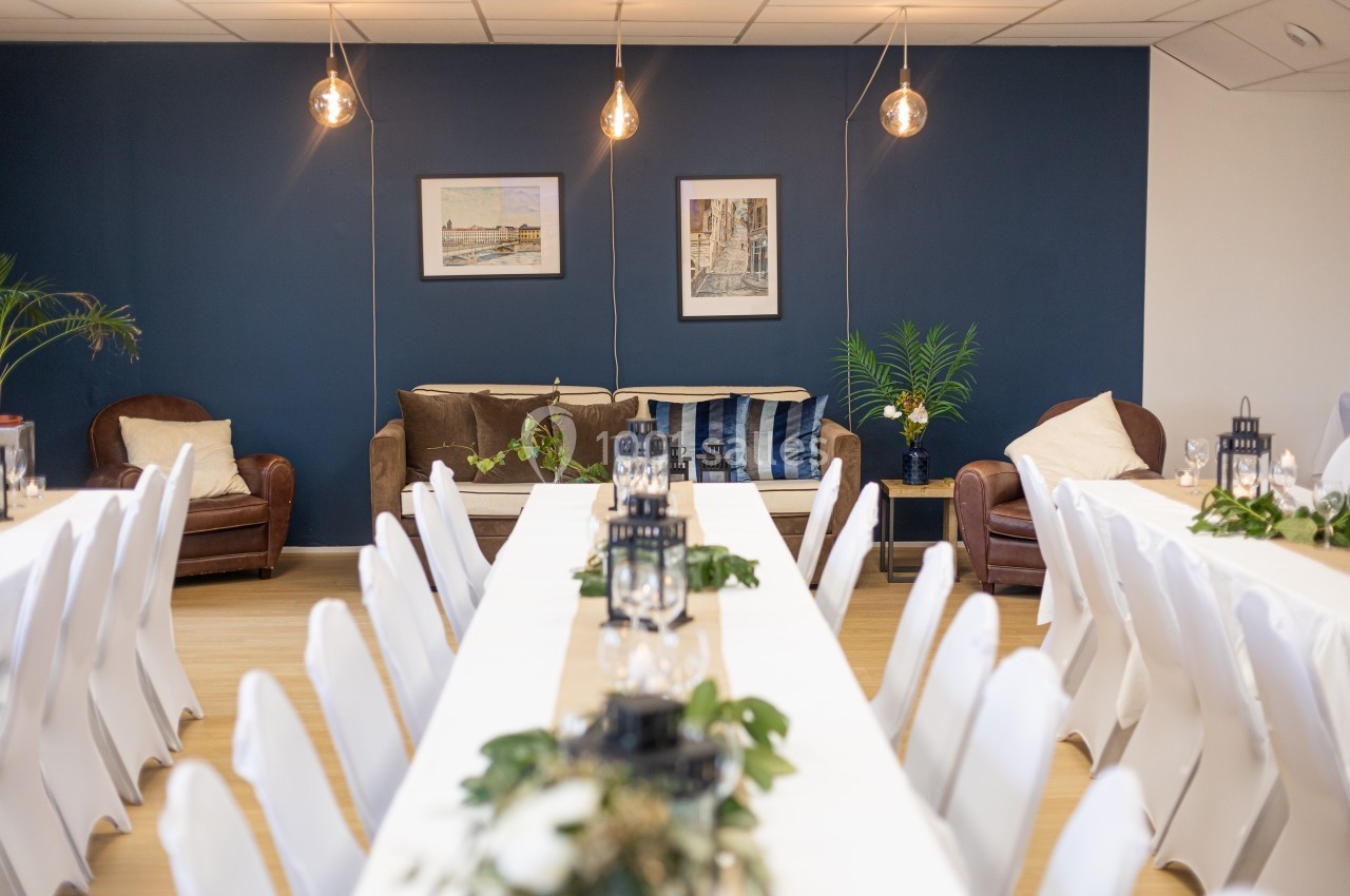 Salle décorée avec des tables blanches alignées, des lanternes, des plantes et un mur bleu en arrière-plan.